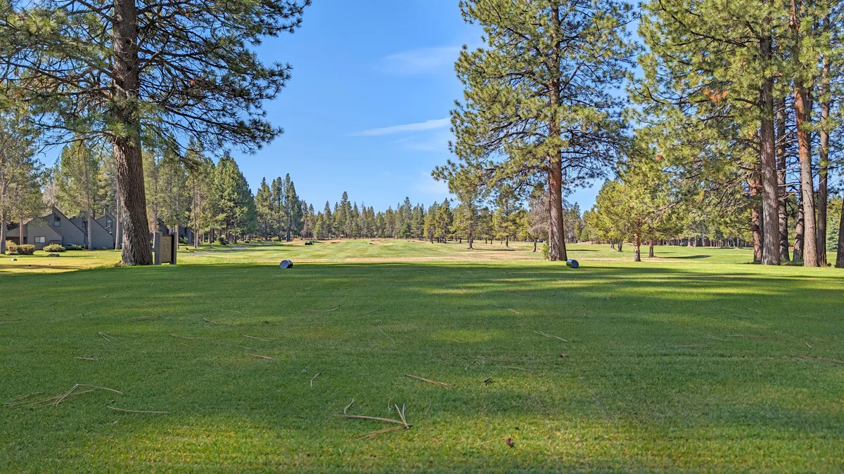 Sweeping view of a lush green golf course, flanked by tall pine trees and stretching into the distance under a clear sky. The layout evokes a sense of spaciousness and connection to nature, ideal for both active and leisurely pursuits.