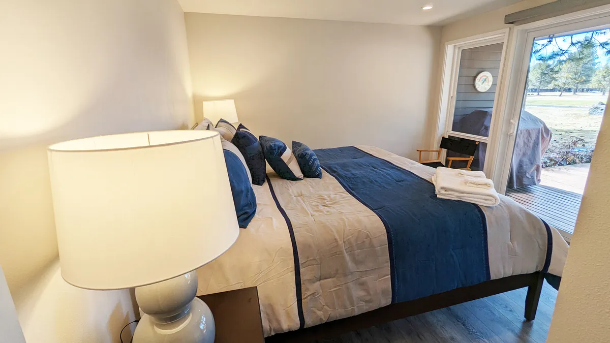 A tranquil and cozy bedroom with a large, inviting bed adorned with decorative pillows. A nightstand with a lamp sits beside the bed, and an open door leads outside, suggesting both comfort and a seamless indoor-outdoor connection.