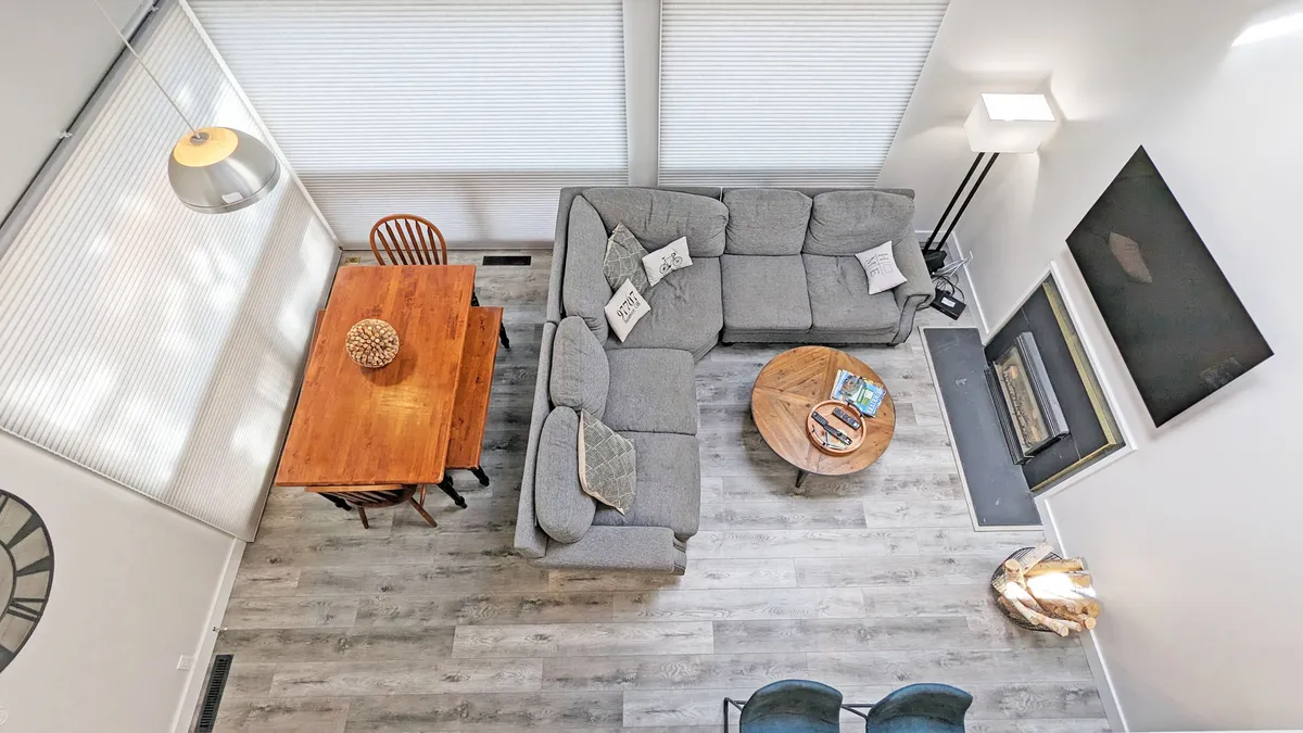This image shows a bright and inviting open living area featuring a plush sofa, a modern dining table, a fireplace, and a stylish light fixture. The space is filled with natural light, and the arrangement encourages relaxation, conversation, and group gatherings.