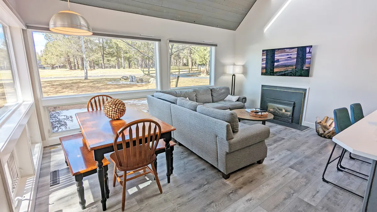 The hero image showcases a bright, inviting living space with expansive windows framing scenic views, a cozy sofa arranged around a modern gas fireplace, an elegant dining table perfect for gatherings, and a large TV. Sunlight pours into the room, highlighting the comfortable, open layout ideal for both relaxation and shared moments.
