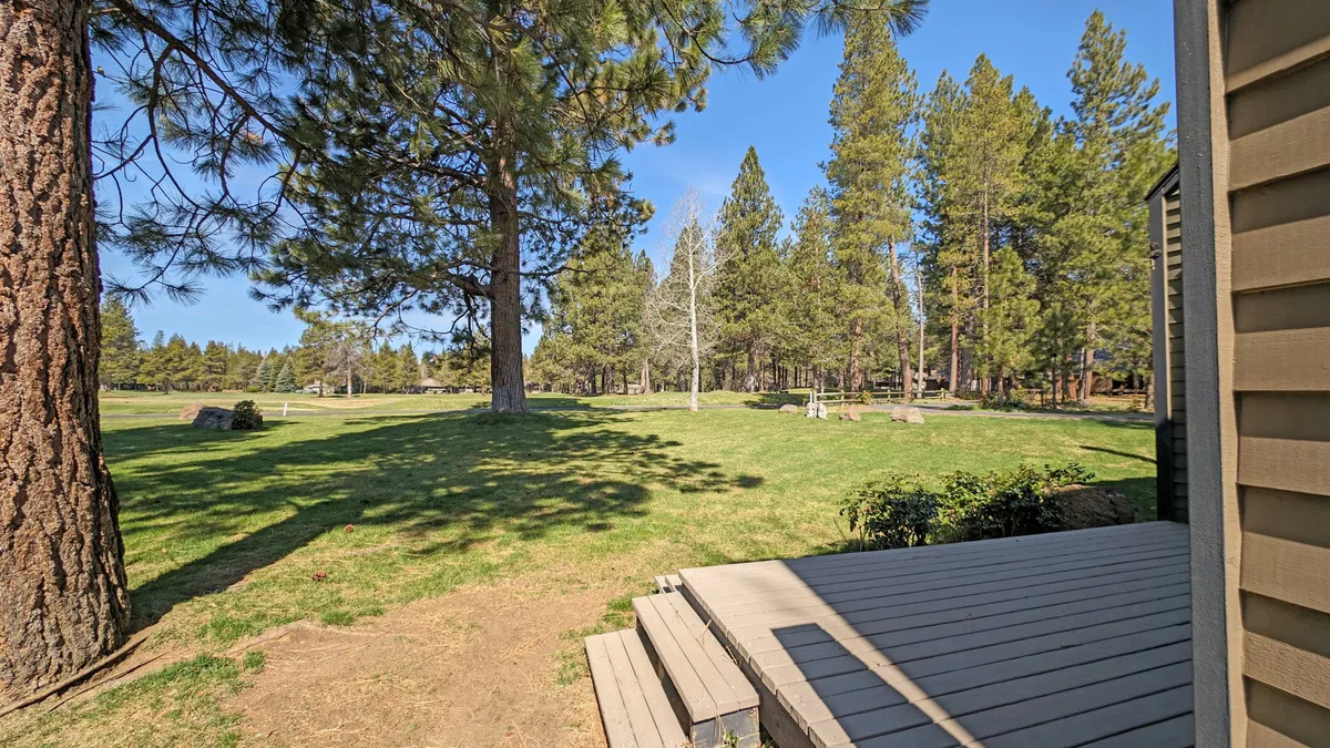 A peaceful exterior shot featuring a spacious lawn bordered by mature trees, with a deck area that invites relaxation and outdoor gatherings. The natural surroundings and open space provide an inviting setting for group activities or quiet contemplation.