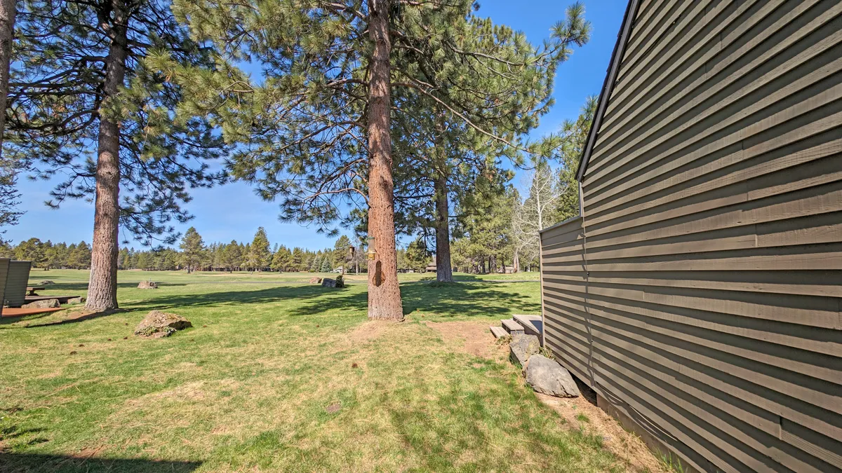 An expansive grassy area shaded by tall pine trees, with a tranquil side view of the building. The scene conveys a sense of space, serenity, and opportunities for relaxing or social gatherings after a day on the course.