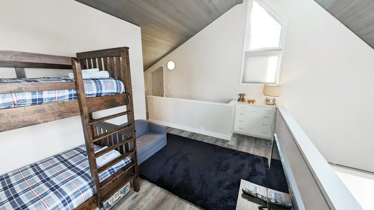 A bright loft space featuring a bunk bed, a blue couch, a nightstand with a lamp, and a window letting in natural light. The playful layout hints at flexibility, perfect for kids or teens, and encourages relaxation, reading, or game time.