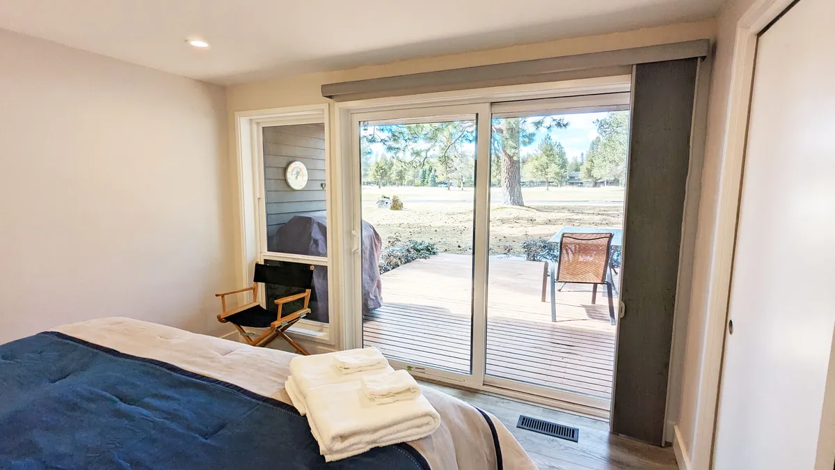 A serene bedroom featuring a plush bed with crisp linens, complemented by sliding glass doors that open directly to an outdoor deck. Natural light streams into the room, enhancing the sense of calm and privacy. A cozy nightstand and soft lighting add to the inviting atmosphere.