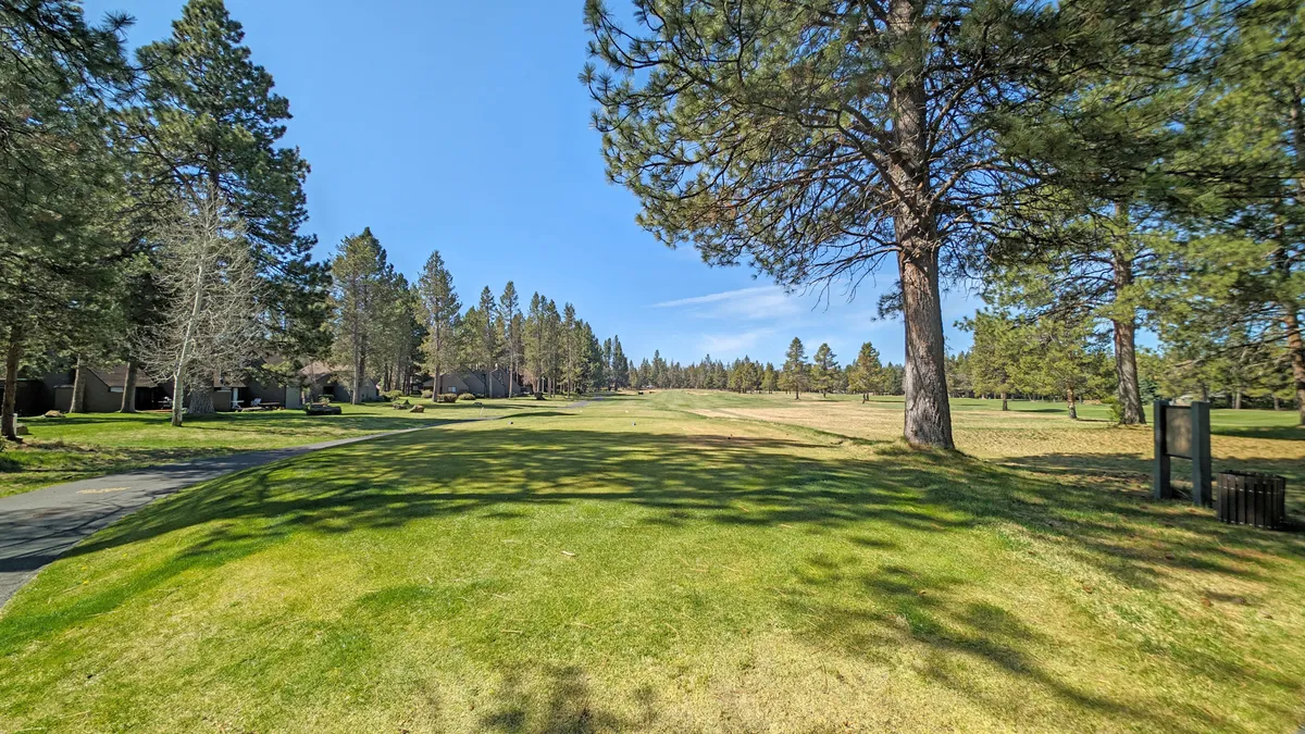 A sunlit view of the lush green golf course, with tall pine trees lining the edges and a clear blue sky overhead. The open fairway invites outdoor activities and highlights the scenic beauty that makes the location special for golf enthusiasts.