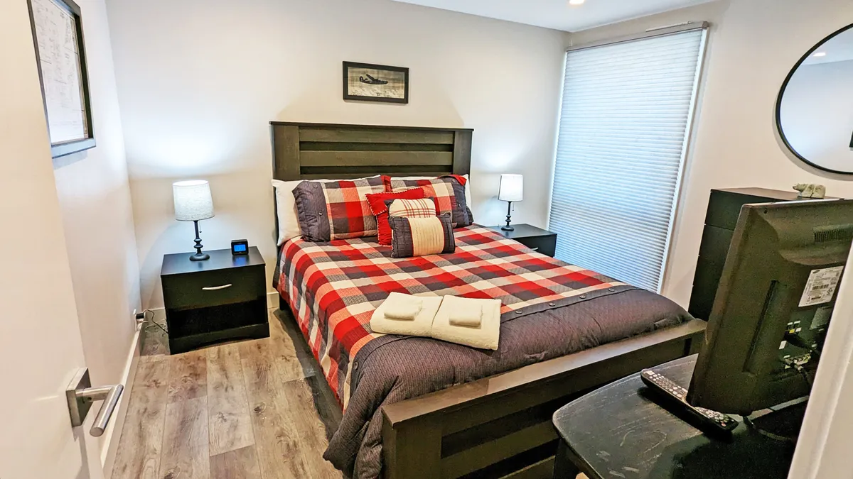 A bedroom with a plaid bedspread, nightstands with lamps, a framed picture above the bed, and a visible television. The space feels warm and inviting, offering a glimpse of the personal retreat awaiting guests, with a focus on cozy relaxation and entertainment.