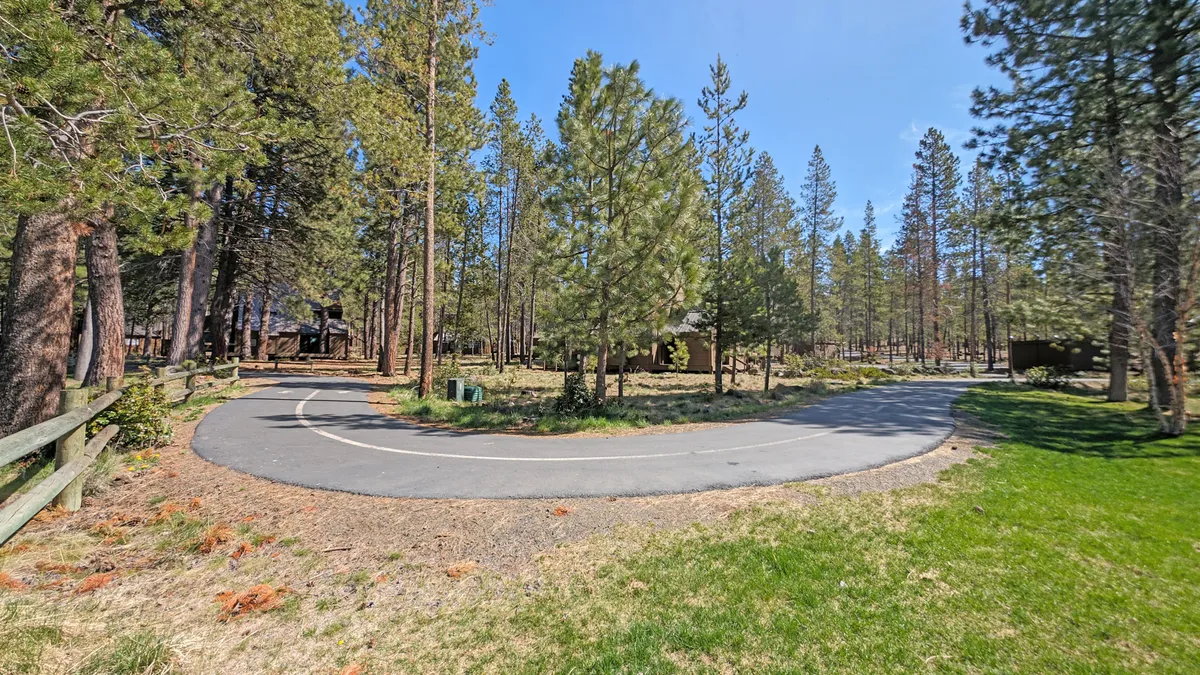 This image captures a winding road flanked by tall trees, with cozy cabins visible in the background. The scene suggests a tranquil, natural setting ideal for biking or setting out on an outdoor adventure.