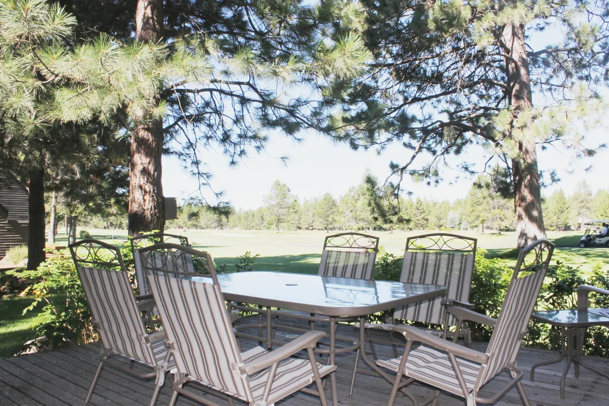 An inviting outdoor dining table and chairs sit on a deck, framed by trees and featuring a clear view of the golf course. The tranquil setting provides a seamless extension from the home into the scenic outdoor environment, ideal for meals with a view.
