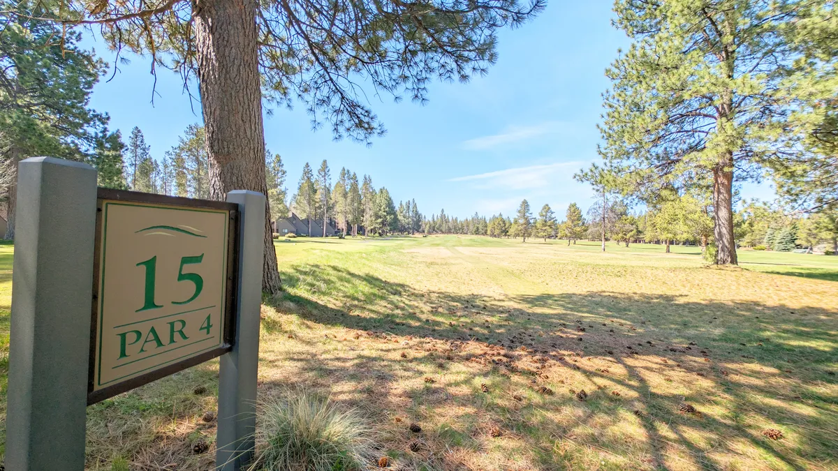 A serene scene of a golf hole sign marking the start of a fairway, bordered by lush green grass and surrounded by tall trees. The open landscape suggests a peaceful retreat perfect for a morning round of golf or a relaxed walk, with plenty of space for group activities.