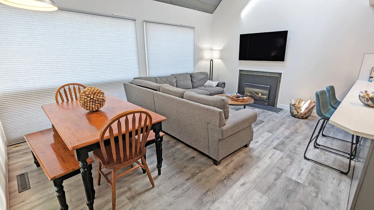 A warm, welcoming interior featuring a comfortable sofa, a modern dining table, a fireplace, and a mounted television. The layout encourages gathering for meals, movies, or sharing stories by the fire.